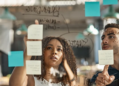 Two people collaborate together standing at a glass whiteboard with post-its and text.